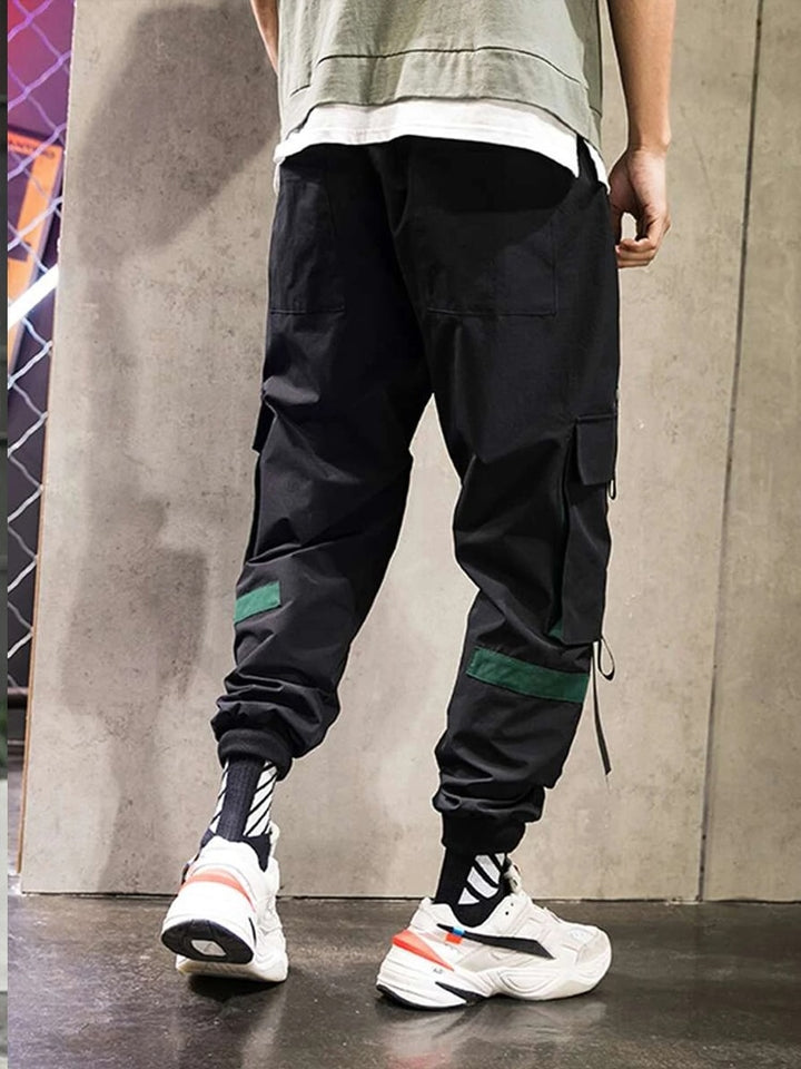 Un homme porte un pantalon cargo noir avec des lanières resserrées aux chevilles. Le pantalon a un style street masculin avec des lanières noires et vertes sur les poches et les jambes. Disponible du M au 5XL, il offre un confort et un maintien accrus grâce à l'élastique à la taille et au cordon. (Cargo District)