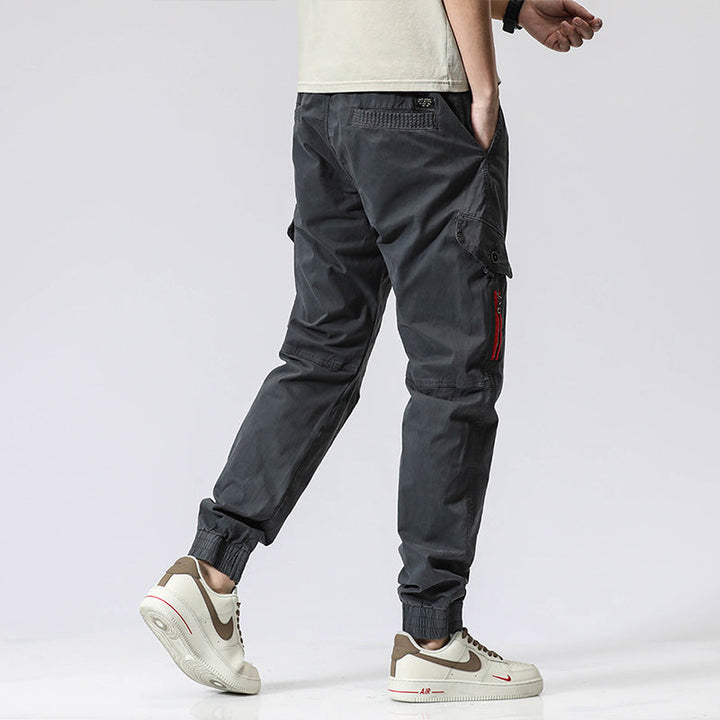 Un homme porte un pantalon cargo gris avec une fermeture rouge. Coupe droite et resserrée à la cheville, ce pantalon est élégant et décontracté. Disponible dans les tailles 28 à 38 (US).