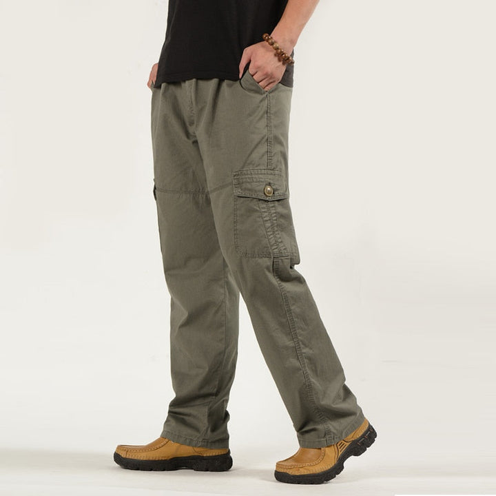 Un homme pose avec un tee-shirt noir, un pantalon cargo coupe droite ample de couleur kaki. Il porte aussi des chaussures camel.