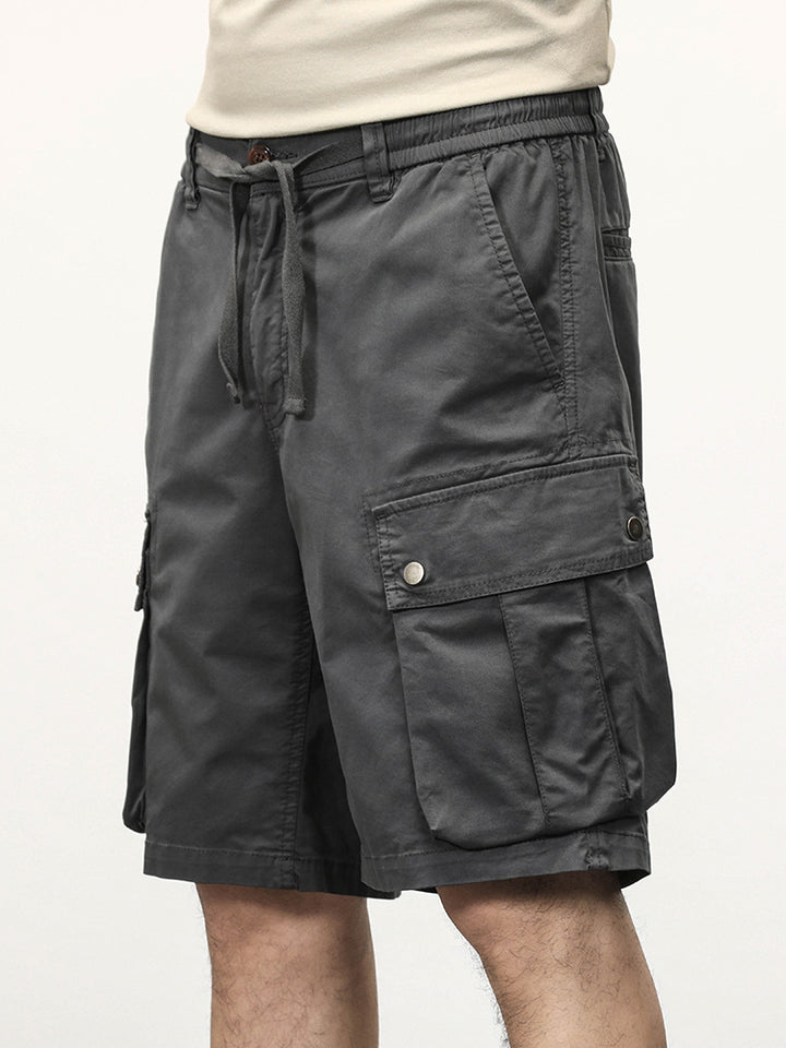 Un homme porte un short cargo gris coupe droite avec des poches latérales à boutons-pression. Le short est fait de 95% coton et 5% spandex pour un confort optimal. Tailles disponibles du 28 au 40.