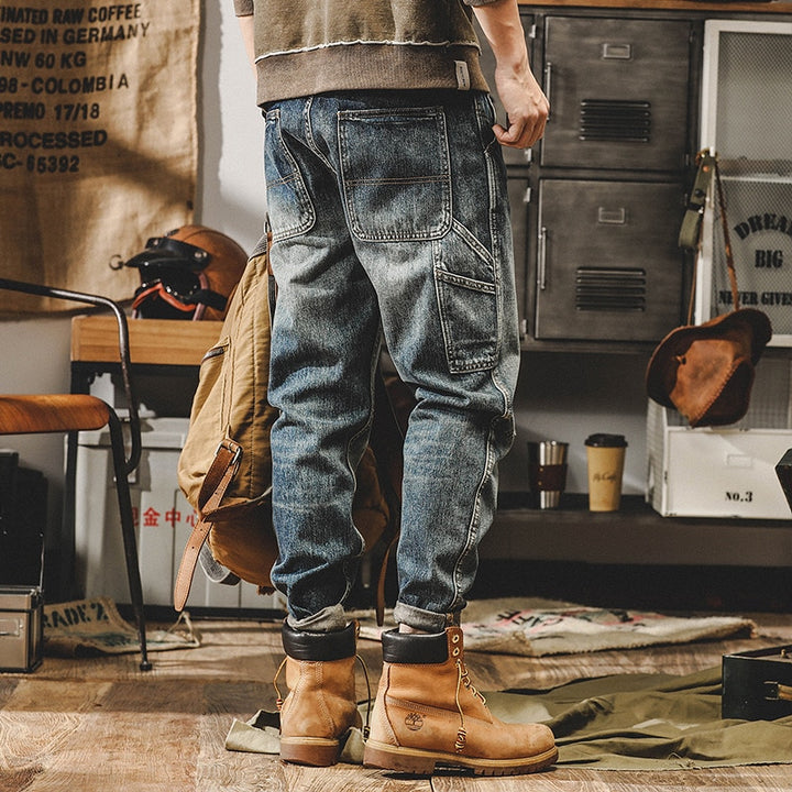 Un homme debout dans une pièce avec un sac à dos, portant un jean cargo délavé coupe droite - Bleu. Les poches latérales ajoutent une touche tendance à votre tenue. Disponible en tailles US du 28 au 40. Conçu en 100% coton pour un confort durable.