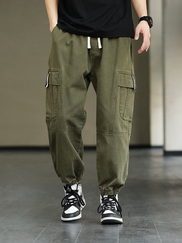 Un homme pose en tee-shirt noir, sneakers noirs et blancs et montre noire. Il porte un pantalon cargo ample kaki avec élastique à la taille et cordon. 