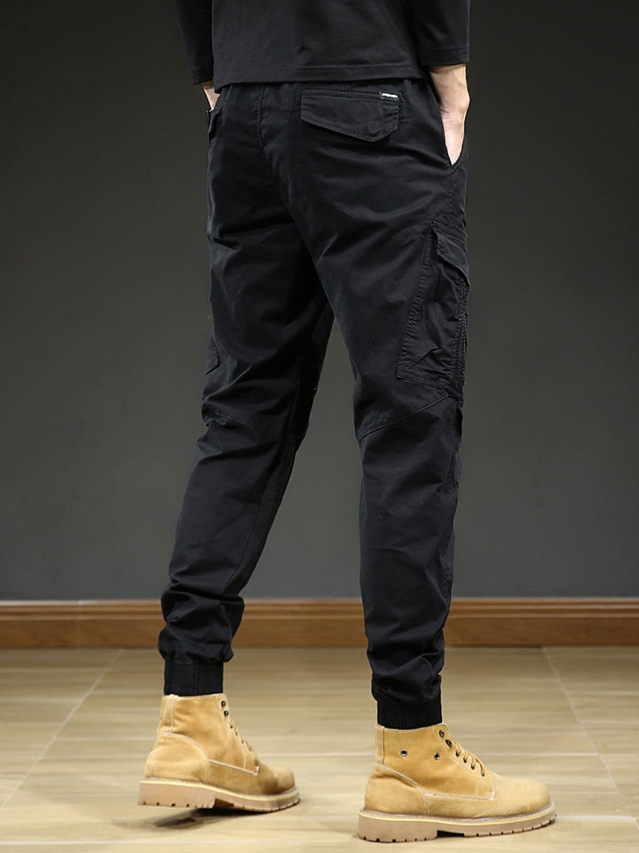 Un homme porte un pantalon cargo slim resserré aux chevilles de couleur noir. Le pantalon est confortable et élégant, avec une taille élastique et un cordon pour un ajustement parfait. La coupe slim et les coutures aux genoux créent un look moderne. Disponible en tailles US 28 à 38. [Cargo District]