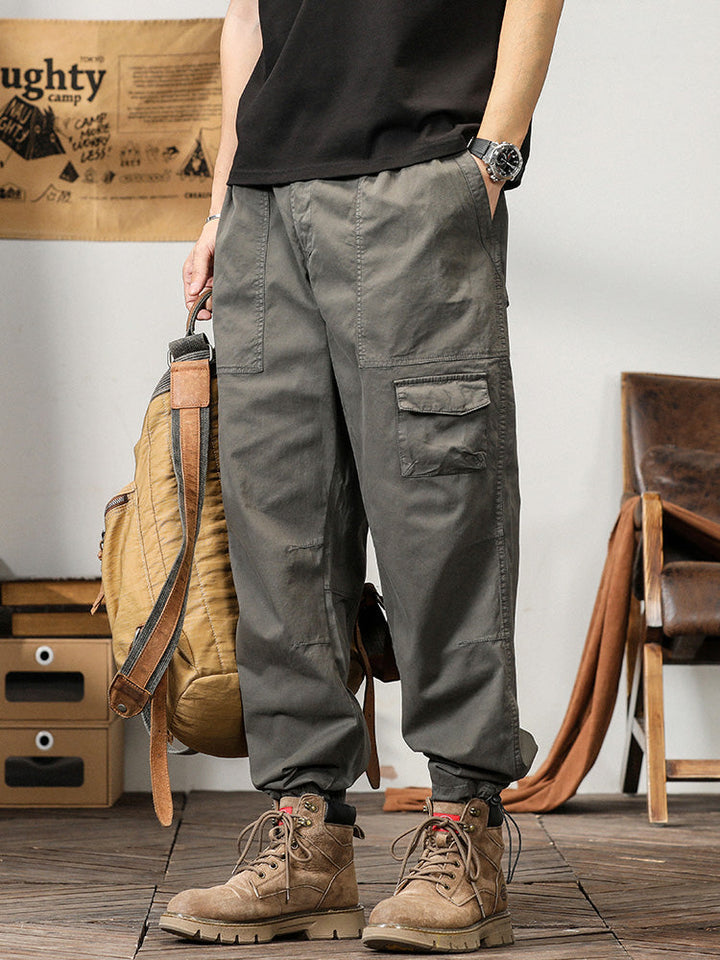 Un homme portant un pantalon cargo léger ample coupe droite gris avec un sac à dos. Le pantalon offre une allure masculine et simple avec des poches fonctionnelles sur les côtés. Confortable et flexible, il est idéal pour l'été. Disponible du M au 3XL.