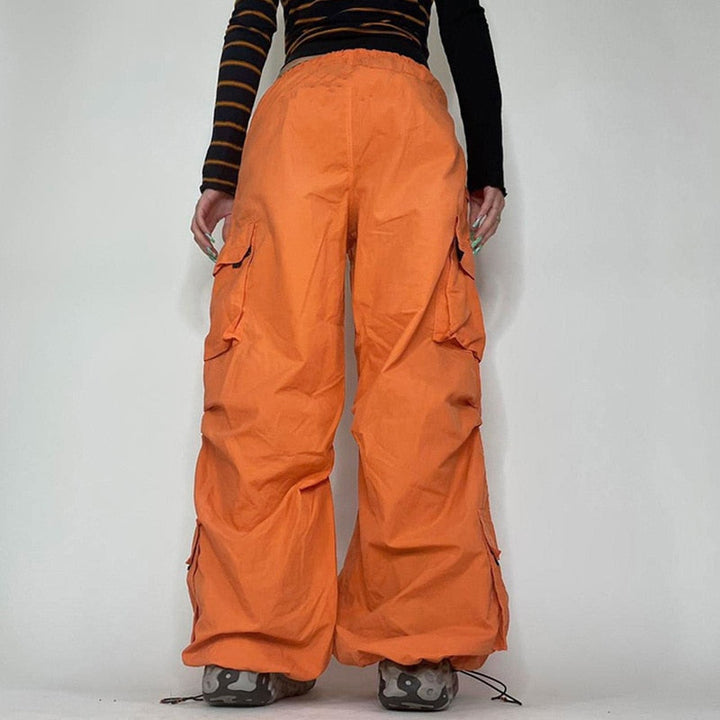 Une personne portant un pantalon orange baggy taille basse pour femme, avec une coupe large façon parachute et des lanières pendantes. Disponible du S au L, ce pantalon offre un confort sans égal et souligne la silhouette. Les chevilles élastiques ajoutent à l'effet baggy, pour un style street féminin unique et audacieux.