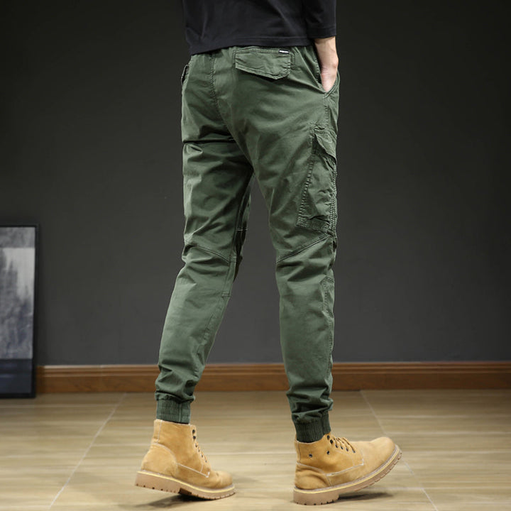 Un homme porte un pantalon cargo slim resserré aux chevilles de couleur kaki avec des bottes marron. Le pantalon est confortable et élégant, avec une coupe slim et des coutures aux genoux. Disponible en tailles US 28 à 38.