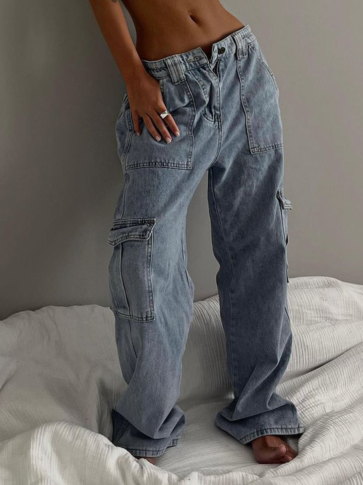 Une femme pose sur un lit blanc avec un jean baggy bleu clair. Elle le porte taille basse et déboutonné. 