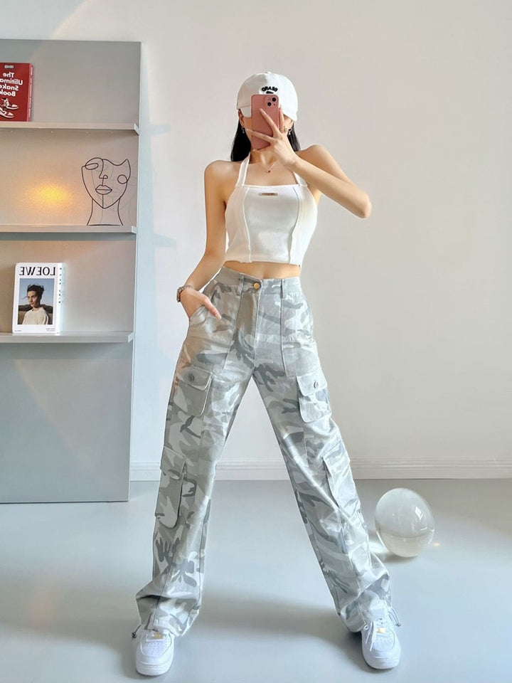 Une femme pose dans un salon aux murs blancs et sol gris. Il y a une bibliothèque dans la pièce. La femme porte une casquette blanche, un top blanc et des baskets blanches. Elle port aussi un pantalon cargo taille haute jambes larges. Le pantalon est gris camo.