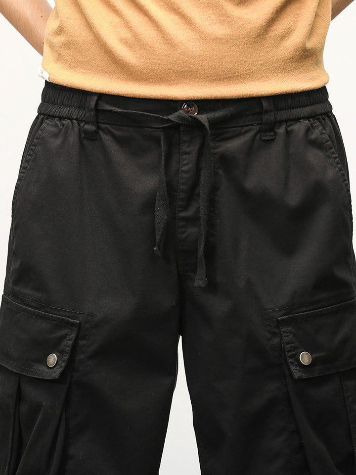 Un homme portant un short cargo noir, coupe droite, avec des poches latérales à boutons-pression. Confortable et pratique, ce short cargo est fabriqué à partir de 95% coton et 5% spandex pour un mélange parfait de confort et d'élasticité. Tailles disponibles du 28 au 40. Parfait pour adopter un style cargo intemporel.