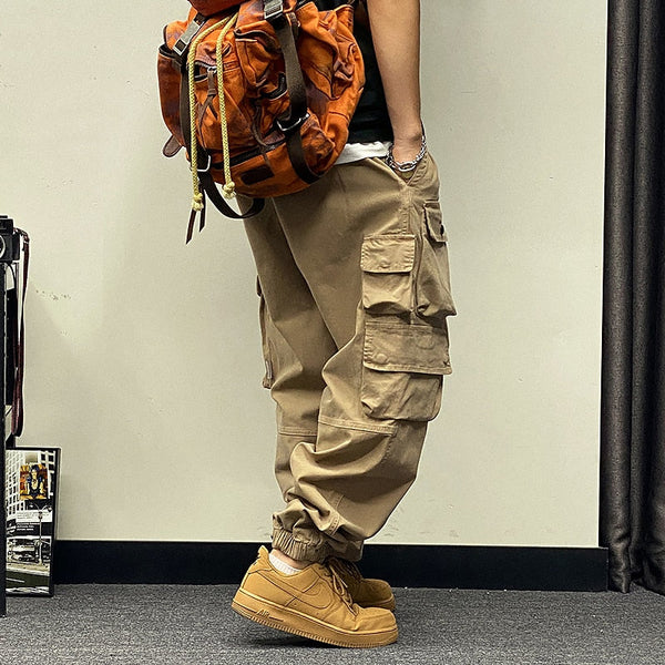 Un homme porte un pantalon baggy à poches asymétriques beige avec des baskets camel, un tee shirt noir et un sac à dos orange. Le sol est gris et le mur beige