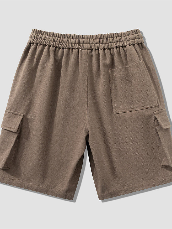 Un homme porte un short cargo marron clair, coupe large, jusqu'aux genoux. Conçu en coton et viscose, il offre confort et flexibilité. Tailles disponibles du M au 8XL.