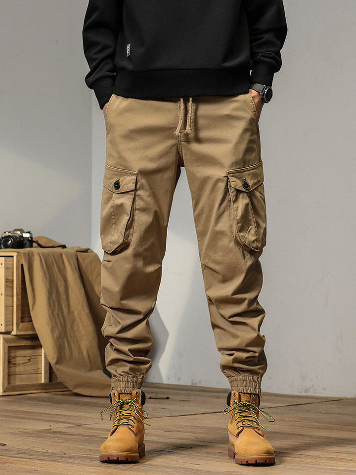 Un homme portant un pantalon cargo beige avec des poches à bouton. Coupe droite et bas resserré aux chevilles. Tailles disponibles du 28 au 38.