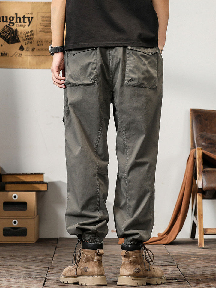 Un homme debout dans une pièce portant un pantalon cargo gris ample coupe droite. Les poches sur les côtés ajoutent une touche fonctionnelle. Tailles disponibles du M au 3XL. Confortable et flexible.