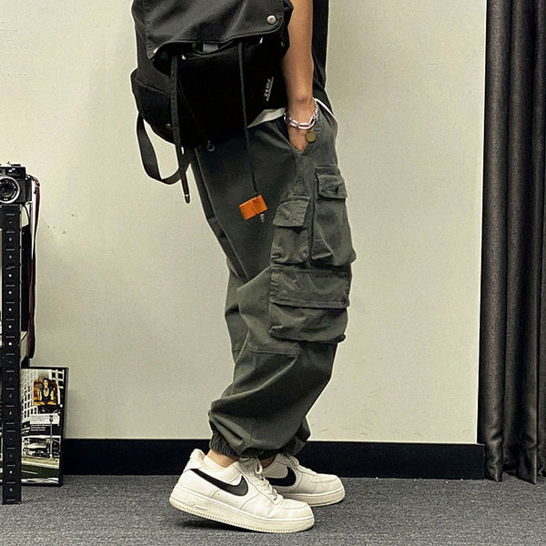 Un homme porte un pantalon baggy à poches asymétriques kaki avec des baskets blanches, un tee shirt noir et un sac à dos noir. Le sol est gris et le mur beige.