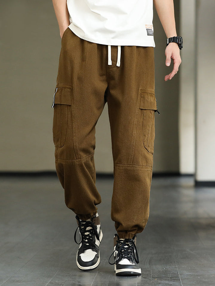 Un homme pose en tee-shirt blanc, sneakers noir, marron et blanche. Il porte un pantalon cargo ample, à taille élastique et cordon blanc. Le pantalon cargo est marron. 