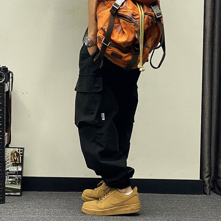 Un homme porte un pantalon baggy à poches asymétriques noir avec des baskets camel, un tee shirt noir et un sac à dos orange. Le sol est gris et le mur beige