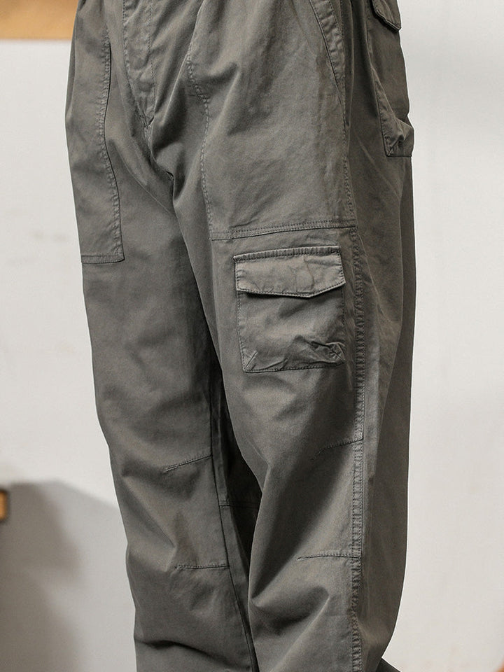 Un homme porte un pantalon cargo léger ample coupe droite gris avec des poches fonctionnelles. Parfait pour l'été, ce pantalon offre confort et maintien grâce à sa taille élastique et sa fermeture classique. Disponible du M au 3XL.