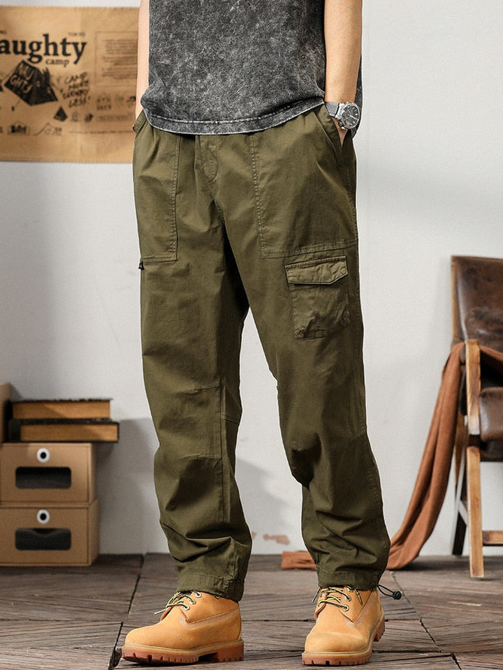 Un homme pose dans un décor industriel avec un tee shirt gris, une montre et des boots marrons. Il porte également un pantalon cargo coupe droite ample et léger kaki.