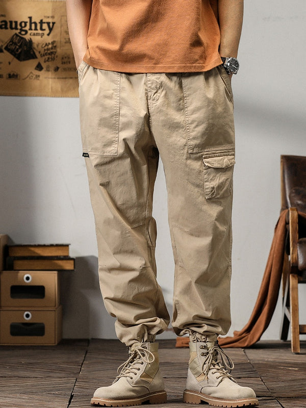 Un homme pose dans un décor industriel avec un tee shirt orange, une montre et des boots beiges. Il porte également un pantalon cargo coupe droite ample et léger beige.