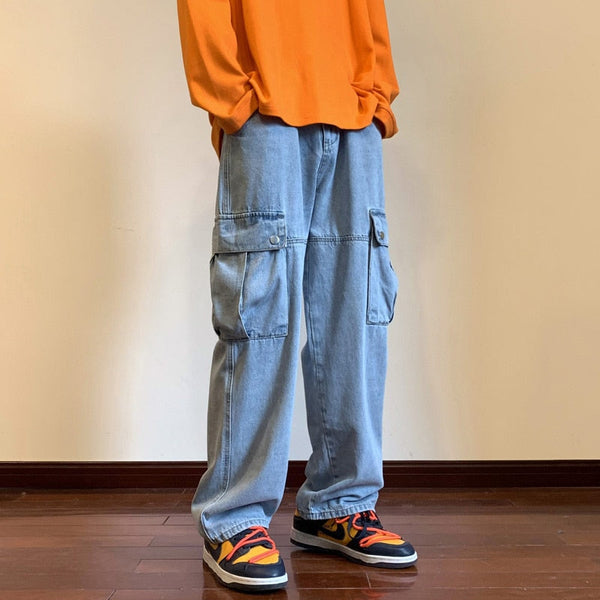 Un homme pose dans une pièce vide aux murs beige et parquet au sol. Il porte un pull orange, des basket noires et oranges ainsi qu'un jean cargo baggy bleu clair.