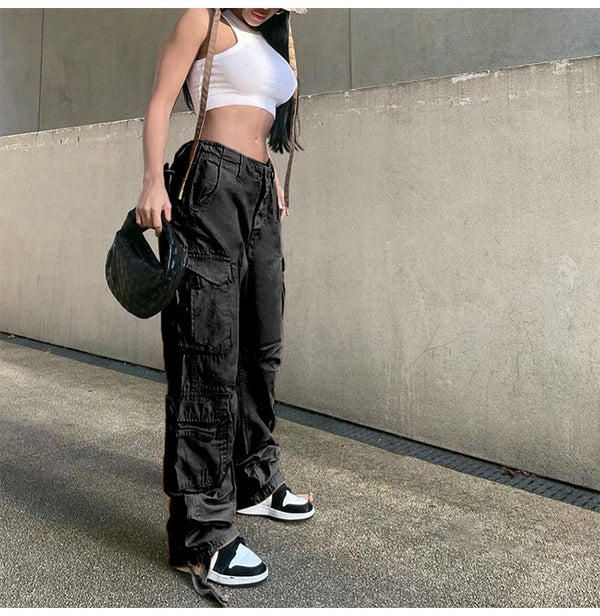 Une femme pose dans la rue avec un pantalon cargo taille haute noir, des baskets noires et blanches, un crop top blanc et un sac à la main. 