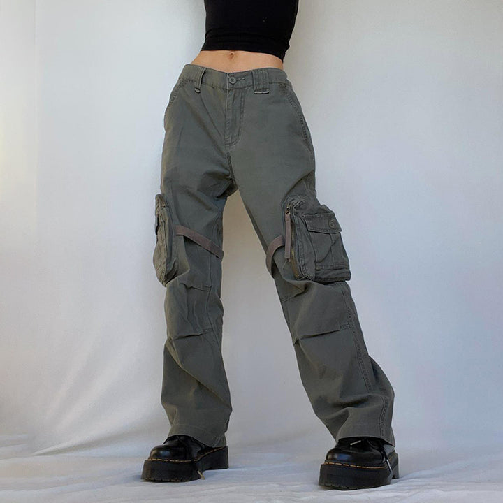 Une personne portant un pantalon cargo large à lanières autour des cuisses kaki pour femme. Le pantalon affiche un look urbain audacieux avec des lanières cousues aux poches latérales, s'enroulant autour de la jambe, ajoutant une touche militaire. Disponible du S au L.