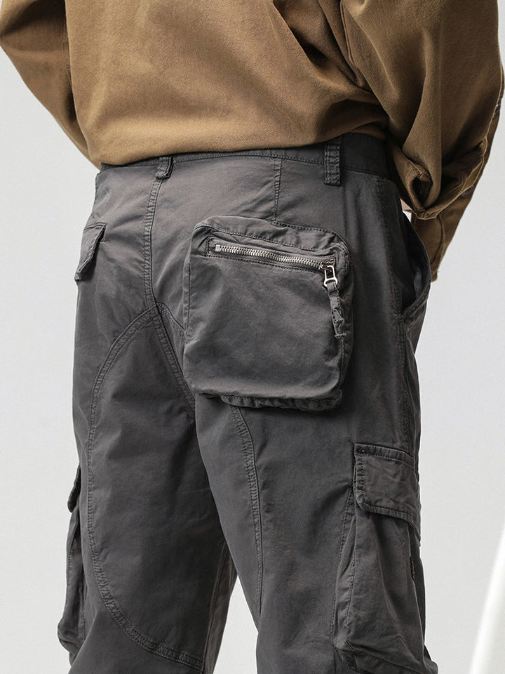 Un homme porte un pantalon cargo ample coupe droite gris avec des poches sur les côtés. Le pantalon est en coton et spandex, offrant confort et flexibilité. Les chevilles peuvent être resserrées avec un lien pour un look sportif. Disponible en tailles du 28 au 38 (US).