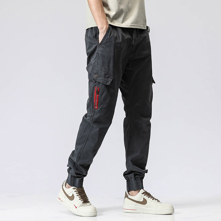 Un homme pose sur fond blanc dans un tee shirt beige, baskets blanches avec une montre et un pantalon cargo coupe droite resserré aux chevilles avec fermeture éclair rouge. Le pantalon est gris.