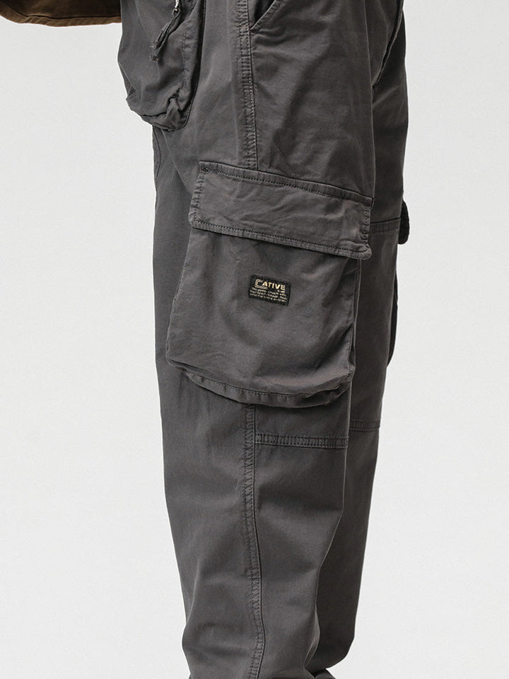 Un homme porte un pantalon cargo gris ample avec poches sur les côtés. Fermeture par bouton et fermeture éclair. Coupe droite, jambe large resserrable aux chevilles. Disponible du 28 au 38 (US). Matières : 97% coton et 3% Spandex. Confortable et flexible.