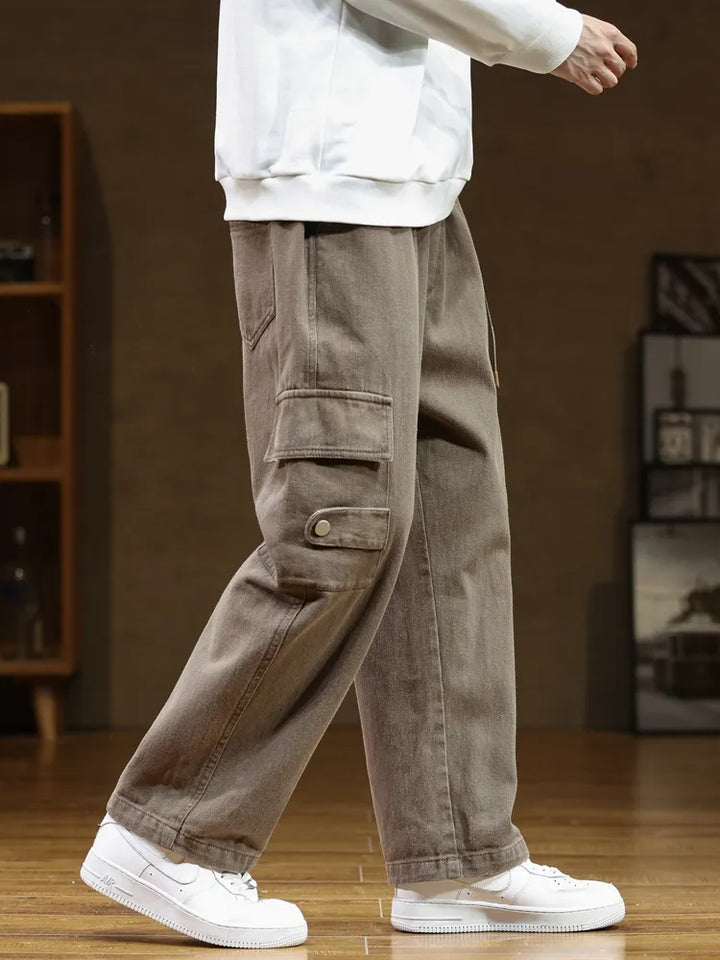 Un homme porte un pantalon cargo ample en coton marron. Ce pantalon résistant offre une liberté de mouvement optimale grâce à sa coupe ample. La ceinture intégrée ajoute une touche utilitaire chic. Disponible en tailles M (FR 36) à 8XL (FR 56).