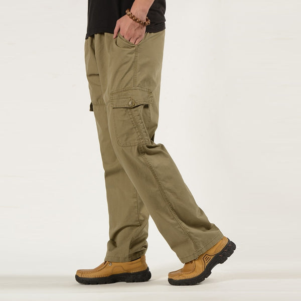 Un homme pose avec un tee-shirt noir, un pantalon cargo coupe droite ample de couleur vert olive. Il porte aussi des chaussures camel. 