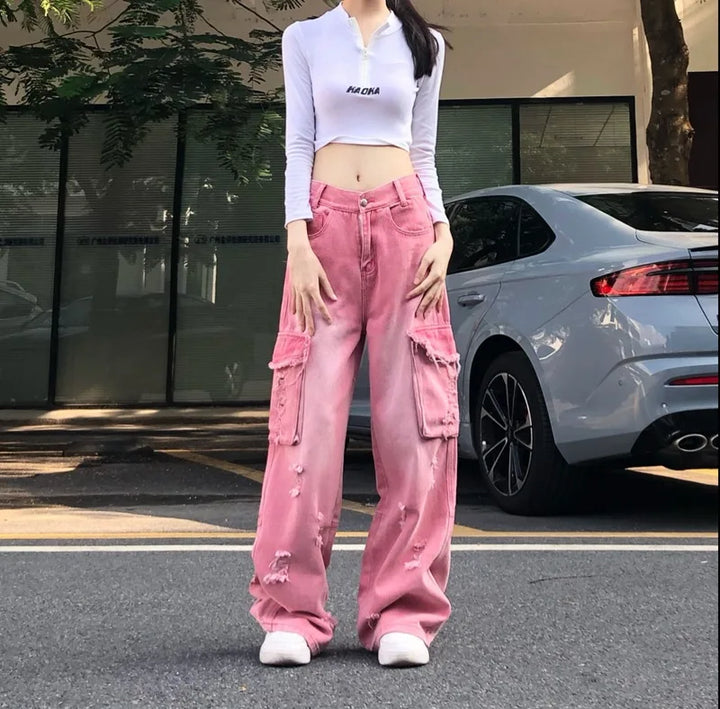 Une femme debout devant une voiture, portant un jean cargo baggy rose délavé déchiré taille haute - Rose - Femme. Un look audacieux des années 2000 avec des déchirures judicieusement placées pour une touche d'insouciance. Disponible en tailles XS à XL. Un véritable atout pour celles qui osent défier les conventions avec style.