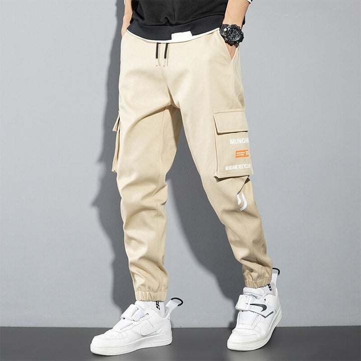 Un homme portant un pantalon cargo jogger beige avec des poches spacieuses et une inscription distincte. Conçu en coton et viscose pour un confort inégalé et une grande durabilité. Disponible du M au 8XL.