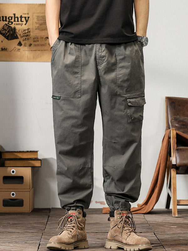 Un homme pose dans un décor industriel avec un tee shirt noir, une montre et des boots marrons. Il porte également un pantalon cargo coupe droite ample et léger gris.