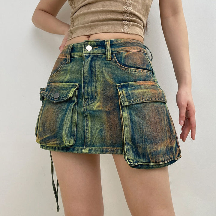 Une femme pose dans une pièce aux murs blancs. Elle porte un tee-shirt beige et une mini-jupe cargo en jean bleu vert multipoches à l'avant.