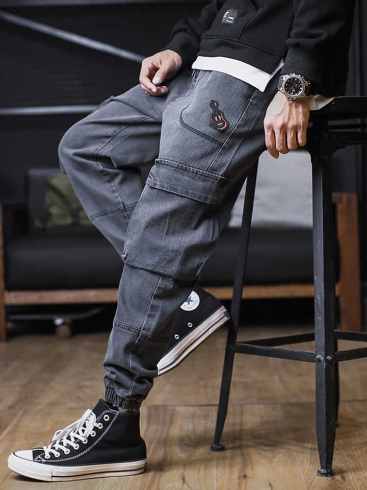 Un homme pose assis sur un tabouret dans un décor industriel. Il porte des converses noires, un pul noir et blanc ainsi qu'une montre. Il porte aussi un jean cargo jogger gris avec un patch guitare sur l'une des poches