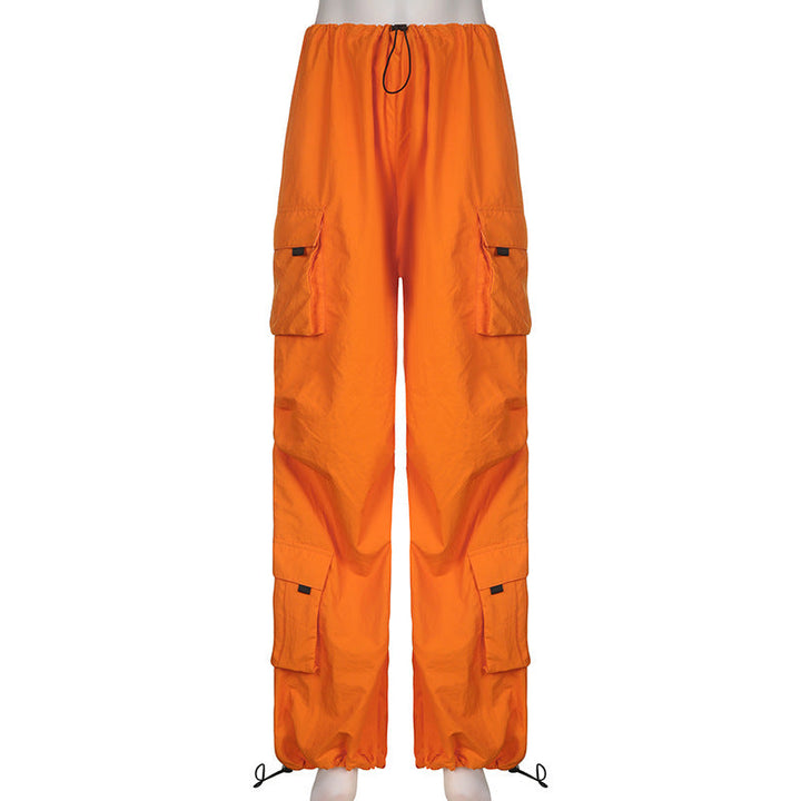 Une personne porte un pantalon orange baggy taille basse pour femme avec des lanières pendantes. La coupe large façon parachute crée une silhouette unique et audacieuse. Disponible du S au L, ce pantalon offre un confort sans égal avec sa taille élastique et ses chevilles élastiques. Matières : polyester, jersey et spandex.