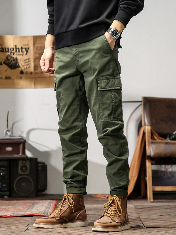 Un homme pose dans un décor industriel avec un sweat noir, une montre et des boots marrons. Il porte également un pantalon cargo coupe droite kaki.