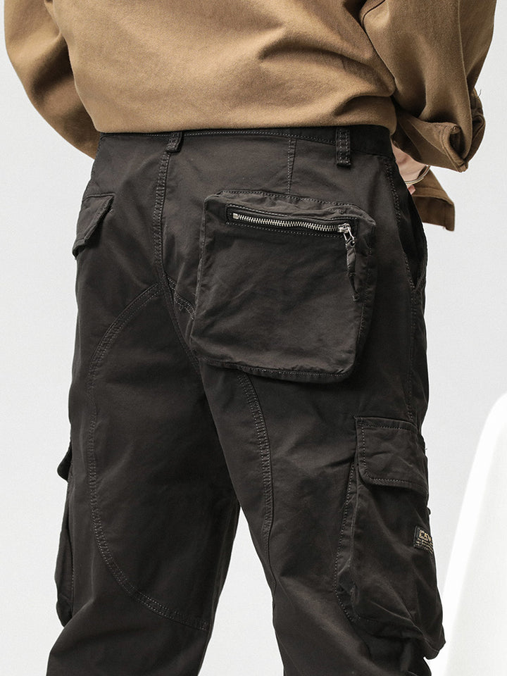 Un homme porte un pantalon cargo noir ample coupe droite avec des poches sur les côtés. Le pantalon est en coton et spandex, offrant confort et flexibilité. Les chevilles peuvent être resserrées avec un lien pour un look sportif ou portées droites pour un look classique. Disponible en tailles US 28-38.