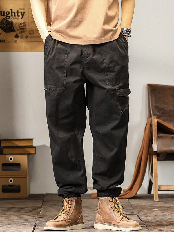 Un homme pose dans un décor industriel avec un tee shirt camel, une montre et des boots marrons. Il porte également un pantalon cargo coupe droite ample et léger noir.