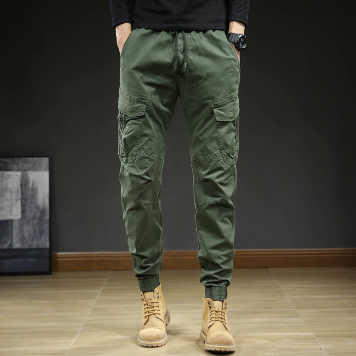 Un homme porte un pantalon cargo slim resserré aux chevilles de couleur kaki avec des bottes brunes. Le pantalon est confortable et élégant, avec une coupe moderne et des coutures aux genoux. Disponible en tailles US 28 à 38.