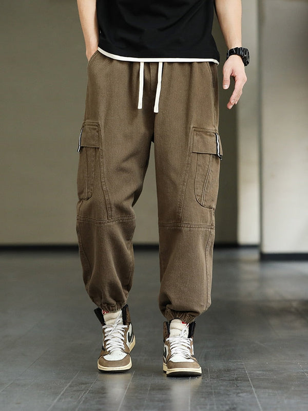 Un homme pose avec un tee-shirt noir et une montre, des sneakers marron et blanches. Il porte un pantalon cargo marron clair avec élastique à la taille et cordon blanc. 