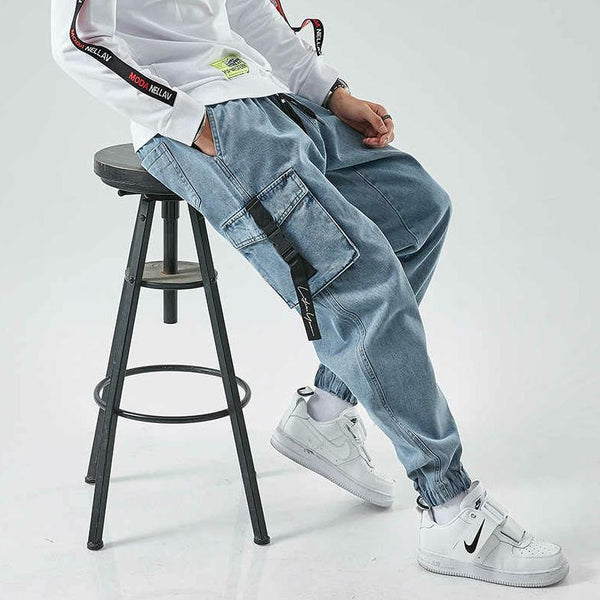 Un homme pose sur un tabouret noir avec un sweat blanc, des baskets blanches et un jean cargo resserré aux chevilles avec des poches à fermeture clip. Le jean est bleu clair. 