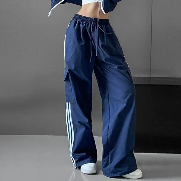 Une femme pose dans une pièce grise. Elle porte des baskets blanches, un crop top blanc, une veste bleu ainsi qu'un pantalon baggy cargo cargo bleu gris avec bandes latérales blanches.