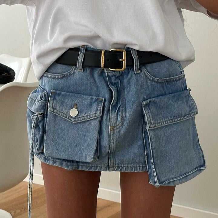 Une femme pose dans une pièce avec parquet au sol et murs blancs. Elle porte un tee-shirt blanc, une ceinture noire à boucle dorée et une mini-jupe cargo en jean multipoches à l'avant. 