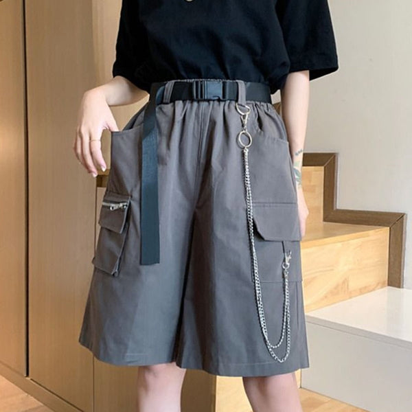 Une femme pose devant des escaliers. Elle porte un tee-shirt noir large ainsi qu'un short long cargo gris avec ceinture noire et chaîne. 