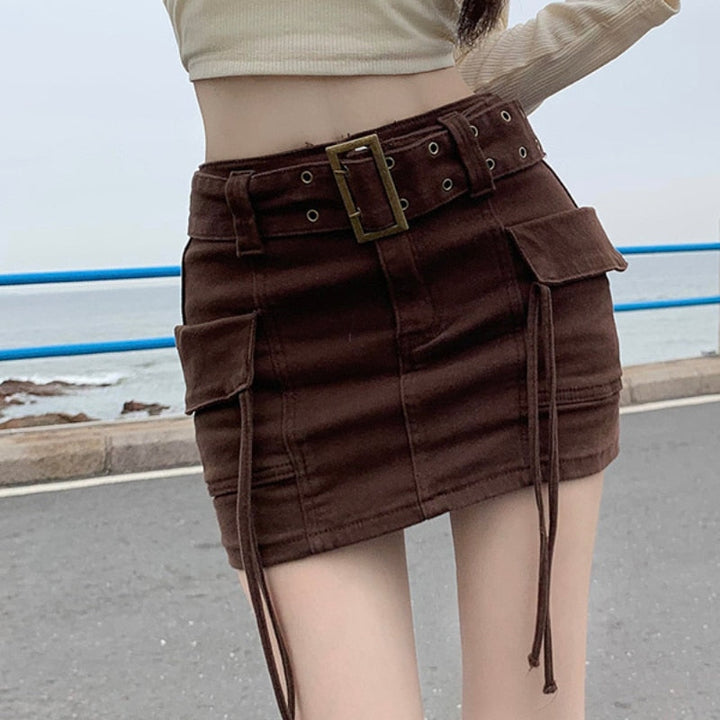 Une femme pose sur une route avec la mer en fond. Elle porte un pull court beige et une jupe taille haute cargo marron avec ceinture et lanières sur les poches latérales.