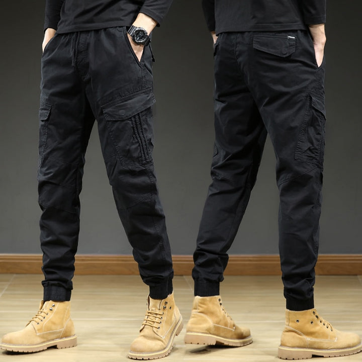 Un homme porte un pantalon cargo slim noir avec des bottes marron. Le pantalon est resserré aux chevilles et possède des coutures aux genoux. Confortable et élégant, il est fait de 95% coton et 5% élasthanne. Tailles disponibles: 28 à 38.