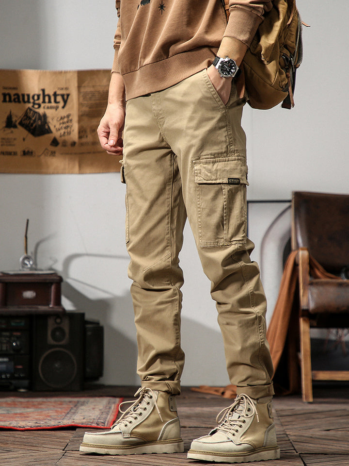 Un homme porte un pantalon cargo beige avec un sac à dos. Le pantalon cargo coupe droite offre un look masculin intemporel avec des poches cargo sur les côtés. Idéal pour une variété d'occasions. Disponible en tailles US 28 à 38.