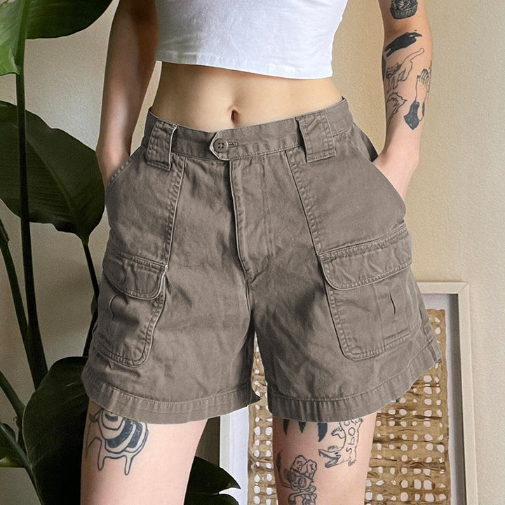 Une femme pose devant un mur beige, avec une plante en fond et un tableau. Elle porte un crop top blanc et un short cargo taille haute gris. Le short est évasé et mi-long. Le mannequin a des tatouages sur le corps.